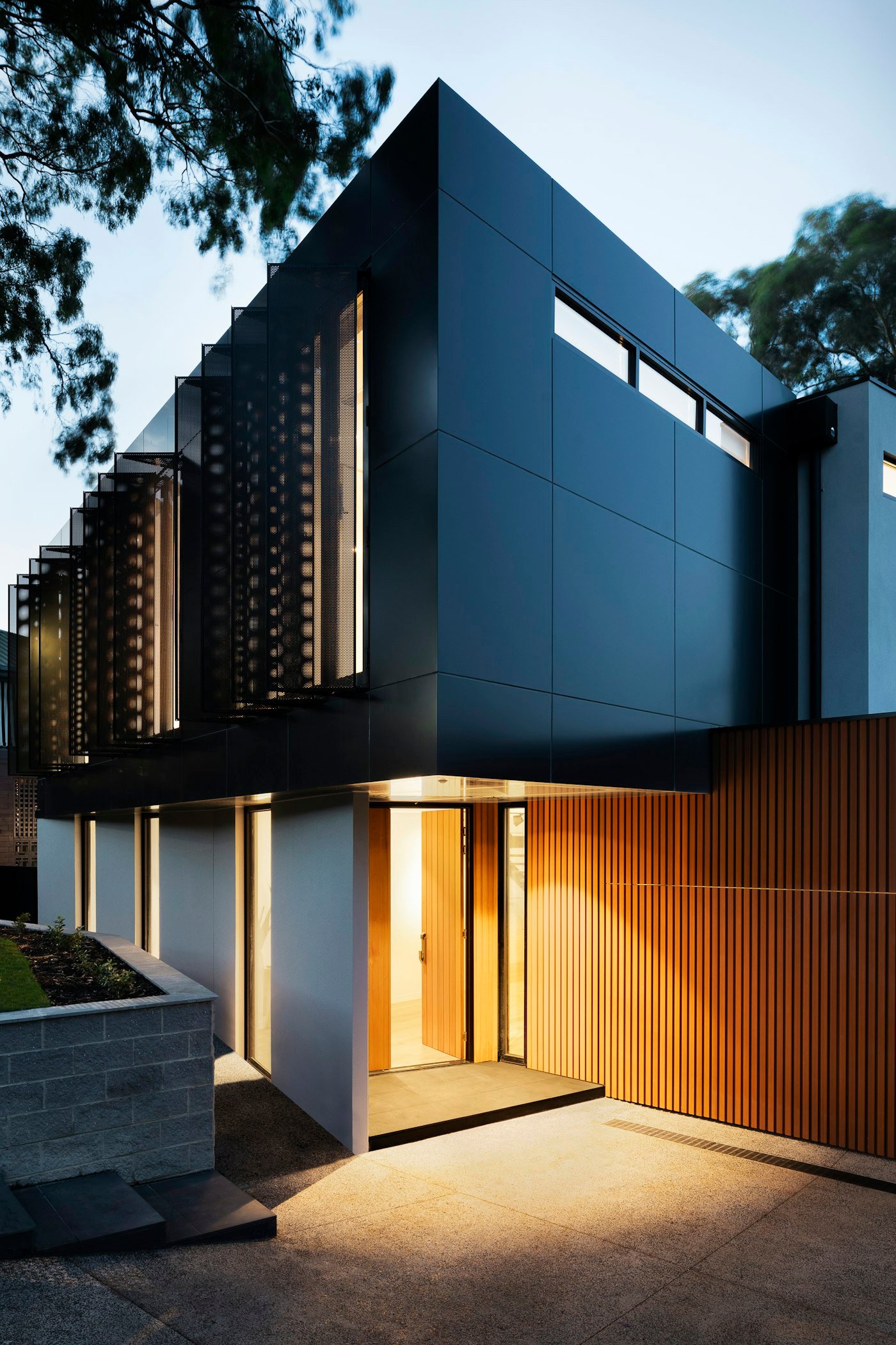 A dark modern house at dusk, with a lit timber front door and timber slats beside the entrance