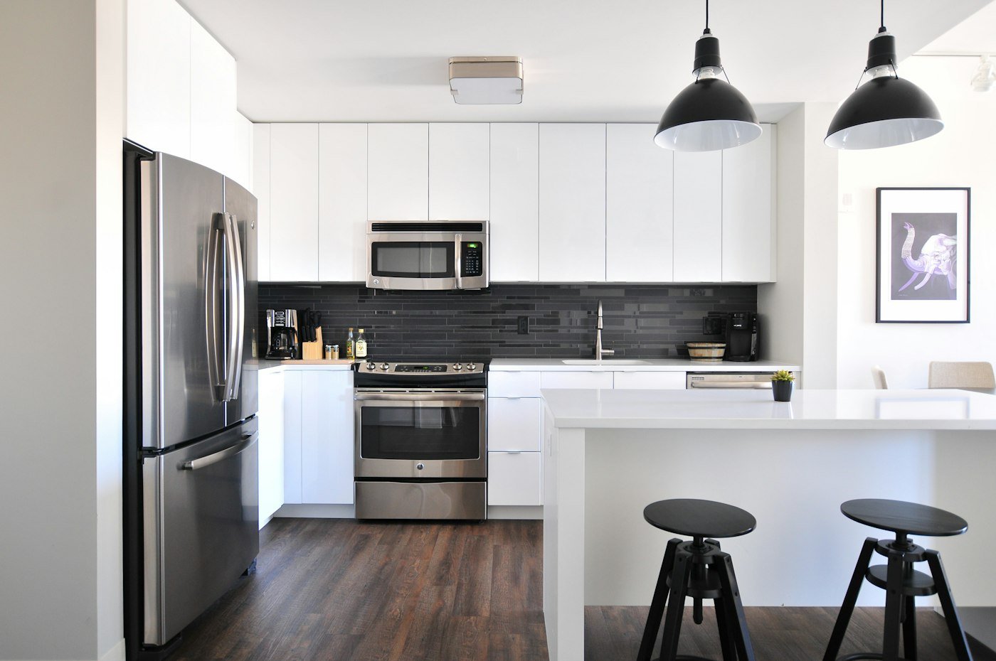A white kitchen with a steel fridge, black tiled splashback, and two black stools at an island